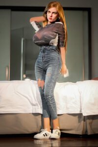 Full-body portrait of a woman in a gray graphic top and ripped jeans, standing beside a bed in a modern room, wearing white sneakers.