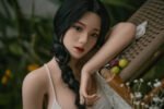 Realistic female doll with braided black hair resting on a woven chair outdoors, beside a small cluster of grapes in a garden.
