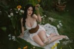 Woman with long dark hair in a white top and brown sash lounging on a lace blanket in a lush garden, framed by green leaves and white daisies.