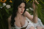Realistic female doll with long black braid, wearing a white bikini top, among green leaves and white flowers.