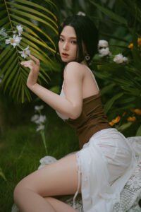 Woman with dark hair wearing a white top and brown belt, kneeling among tropical greenery and white flowers, gazing at the camera.