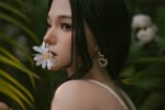 Close-up side profile of a woman with a white daisy held between her lips, wearing a heart-shaped pearl earring, lush greenery in the background