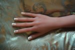 Close-up of a hand with French manicure resting on gold and teal damask-patterned fabric.