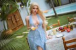 Blonde woman in a plunging light-blue lace dress stands by an outdoor dining table with wine, fruit, and candles near a pool.