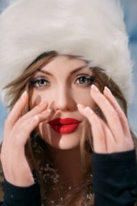Close-up of a woman wearing a white fluffy winter hat, bold red lipstick, and snowy hands framing her face outdoors.