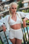 woman with platinum blonde braids wearing a white crop top and shorts, leaning on a railing outdoors on a balcony, wearing hoop earrings and a cross necklace.